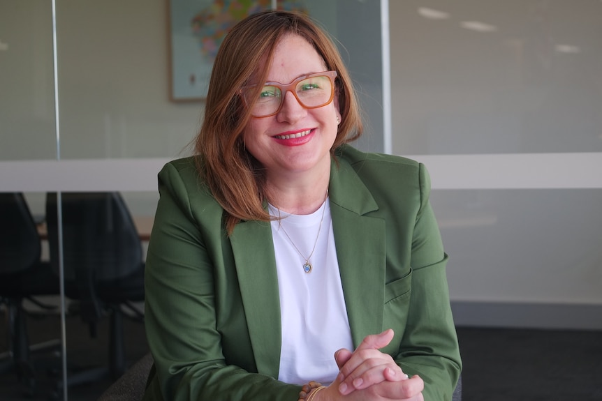 Nicole wears a green blazer and orange-rimmed glasses and smiles with her hands clasped as she sits in a conference room. 